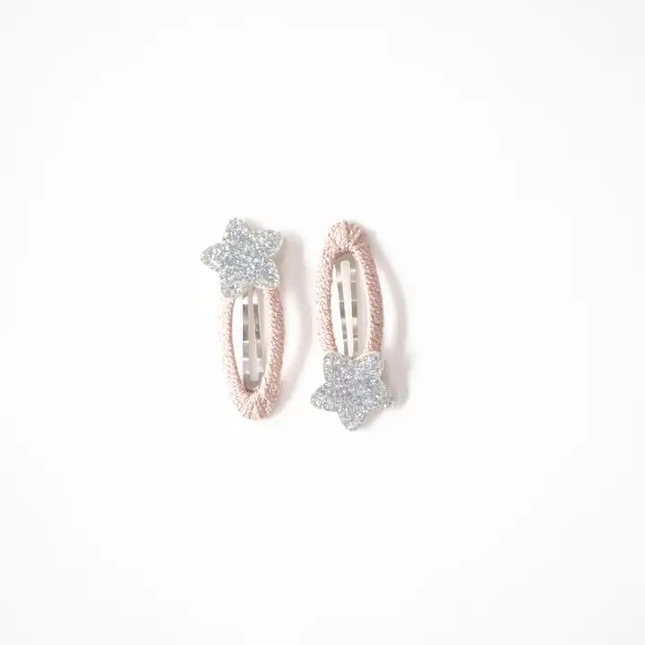 Photo of two kids’ hair clips with silver glitter star accents attached to soft blush-pink oval bases, displayed on a white card with a small gold heart.