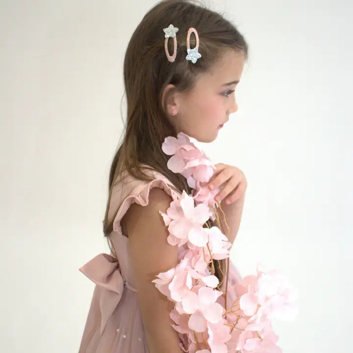 Young girl wearing a pink dress with two silver glitter star hair clips on her side part, holding soft pink flowers.