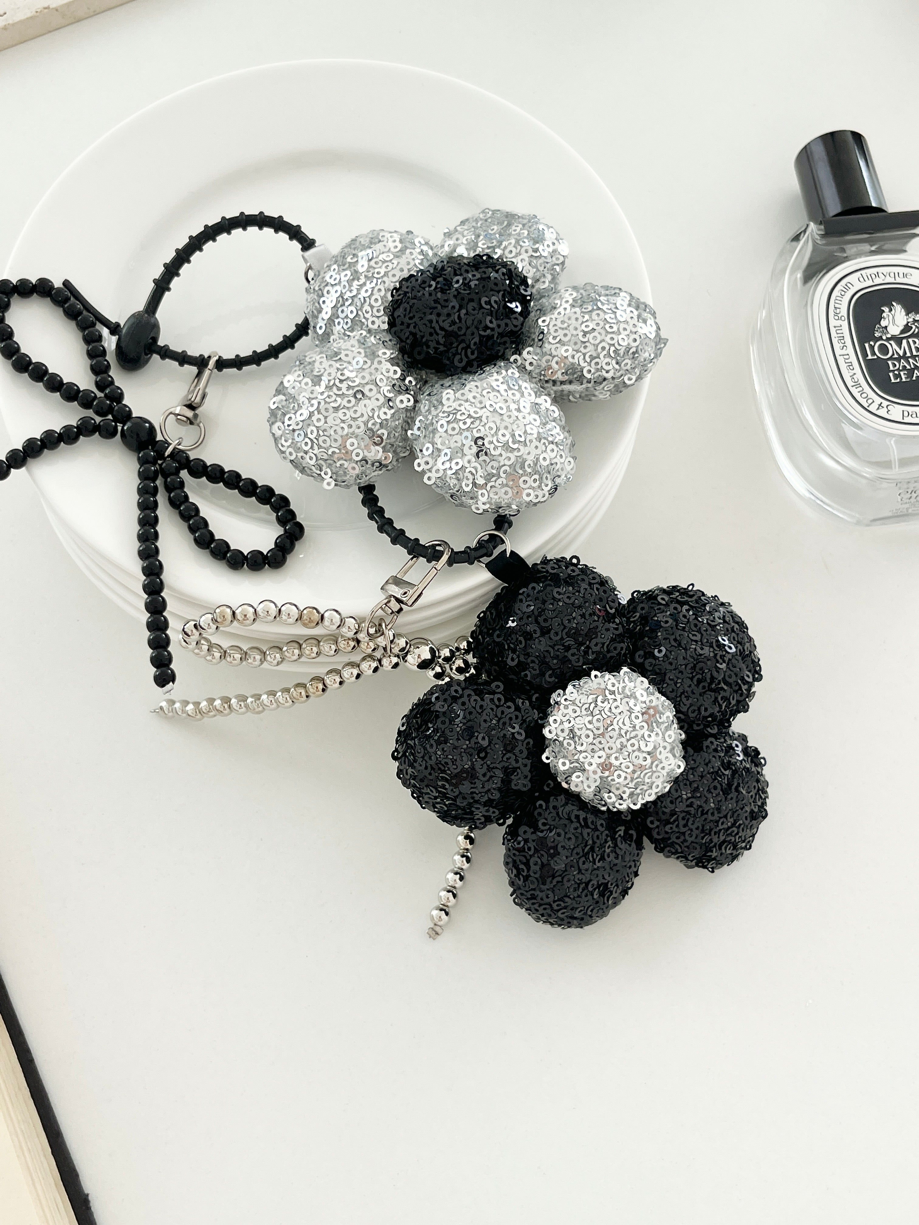 Two sequined flower keychains in black and silver with matching bead bow charms, displayed on a white plates next to perfume bottle.