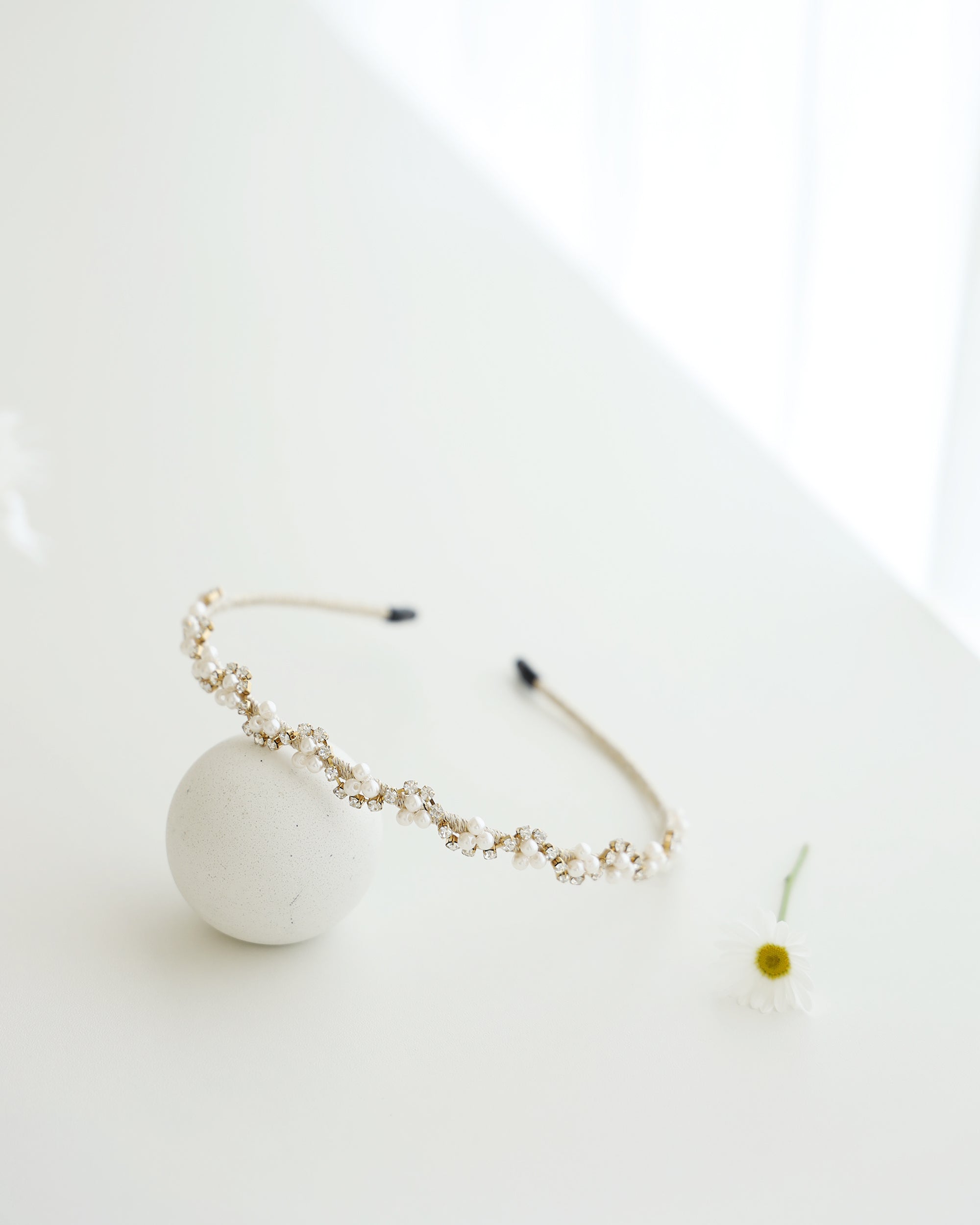 A gold floral headband adorned with small white pearls and crystals, displayed on a white surface with minimalistic props, including a soft white ball.
