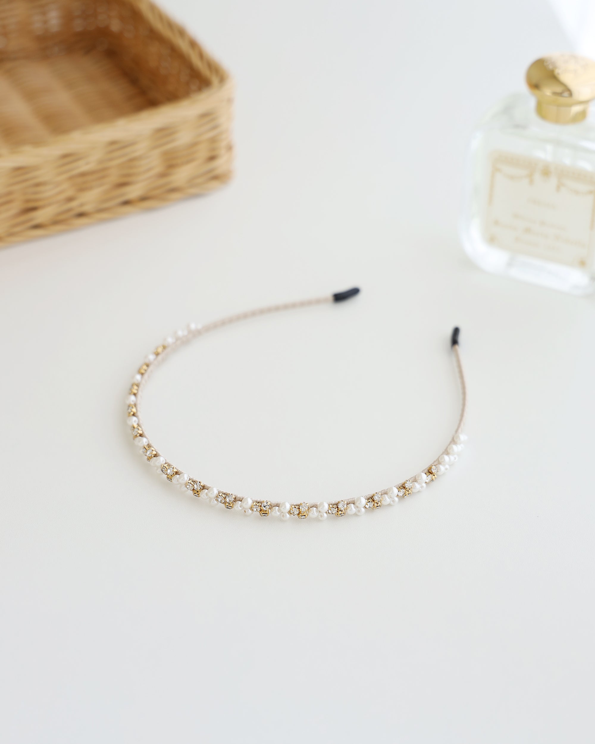 A delicate gold-toned headband featuring small white pearls and sparkling crystals, displayed on white surface with soft natural lighting, perfume bottle and basket.