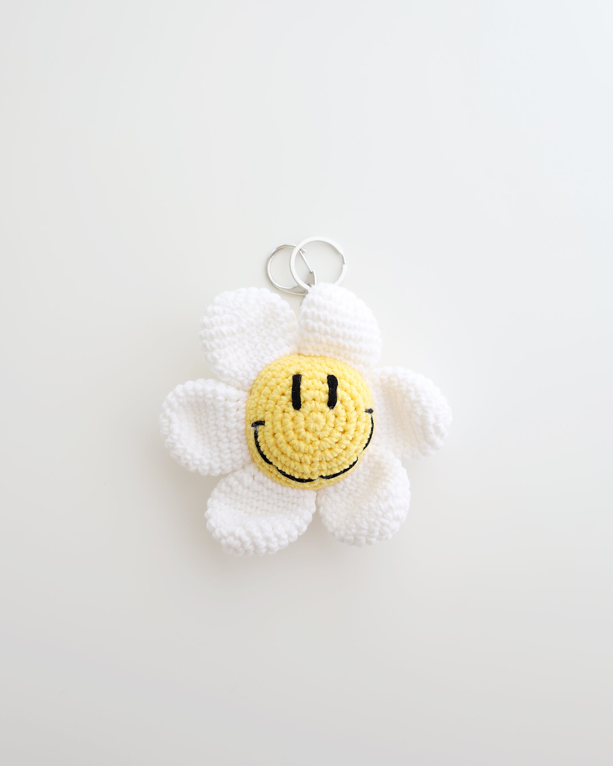 A crocheted flower keychain with soft white petals and a bright yellow smiley face center is displayed. It features a silver keyring at the top for easy attachment to bags, keys, or pouches.