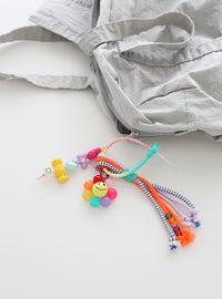 Rainbow-colored beaded flower bag charm with knotted multicolor cords and candy-shaped beads attached to a light gray fabric tote bag.