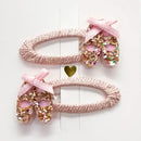 Two pink hair clips decorated with glitter ballet shoes and pink bows on a woven oval base.