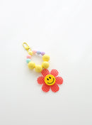 A beaded keychain featuring bright yellow beads and pastel accents, with a red gingham flower charm and a cheerful yellow smiley face center, attached to a yellow clasp.