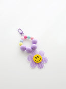 A beaded keychain featuring bright soft purple beads and pastel accents, with a soft purple gingham flower charm and a cheerful yellow smiley face center, attached to a soft purple clasp.