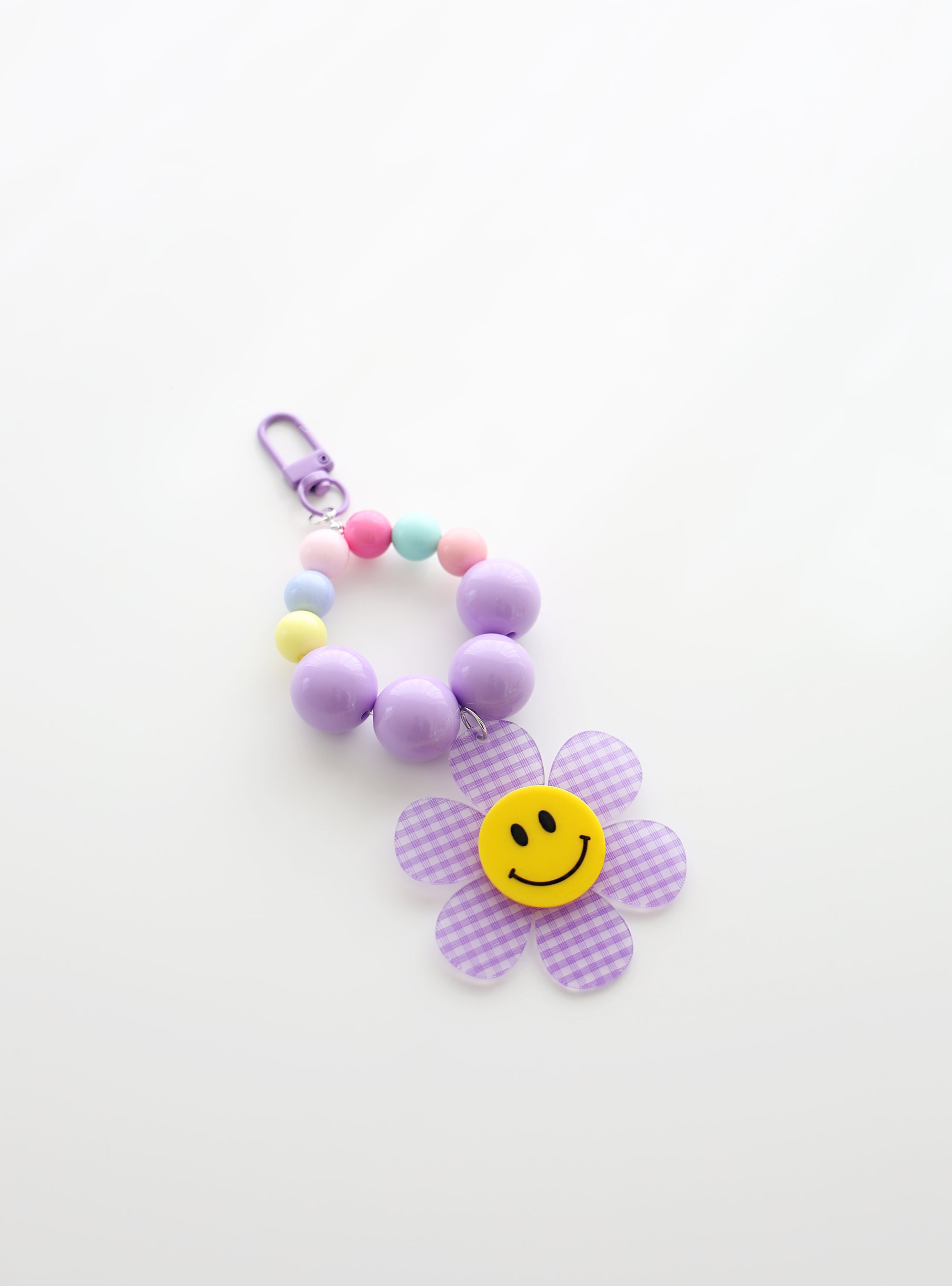 A beaded keychain featuring bright soft purple beads and pastel accents, with a soft purple gingham flower charm and a cheerful yellow smiley face center, attached to a soft purple clasp.