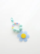 A beaded keychain featuring bright soft sky blue beads and pastel accents, with a soft sky blue gingham flower charm and a cheerful yellow smiley face center, attached to a soft sky blue clasp.