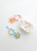 Two colorful beaded keychains with gingham flower smiley charms — one in pink and one in sky blue — shown  attached to or next to a small quilted pouch on a white background.