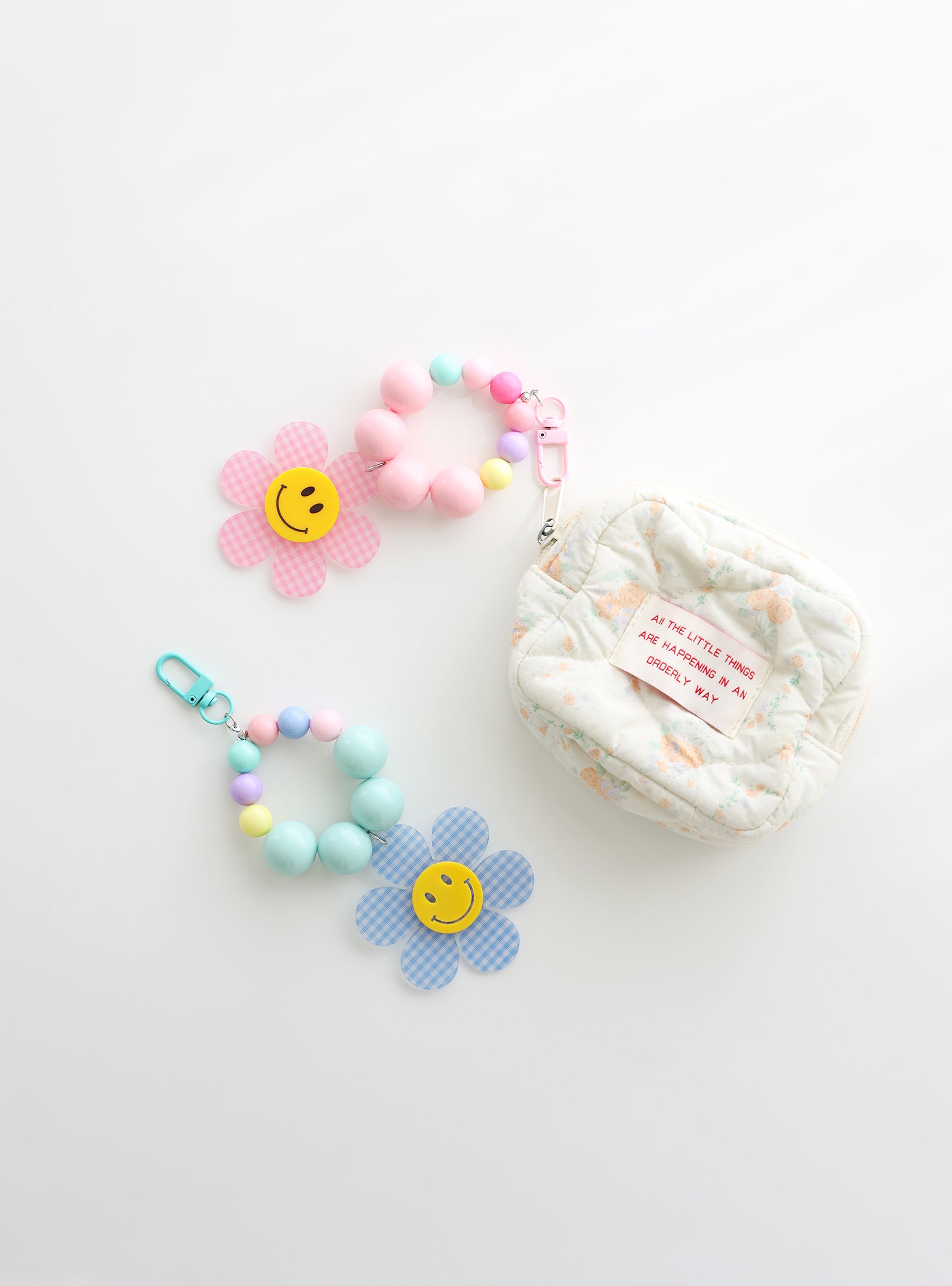 Two colorful beaded keychains with gingham flower smiley charms — one in pink and one in sky blue — shown  attached to or next to a small quilted pouch on a white background.