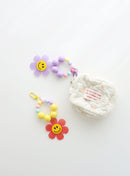Two colorful beaded keychains with gingham flower smiley charms — one in purple and one in red — shown attached to or next to a small quilted pouch on a white background.