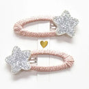Photo of two kids’ hair clips with silver glitter star accents attached to soft blush-pink oval bases, displayed on a white card with a small gold heart.