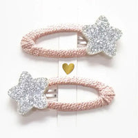 Photo of two kids’ hair clips with silver glitter star accents attached to soft blush-pink oval bases, displayed on a white card with a small gold heart.
