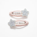 Photo of two kids’ hair clips with silver glitter star accents attached to soft blush-pink oval bases, displayed on a white card with a small gold heart.