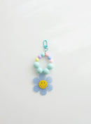A beaded keychain featuring bright soft sky blue beads and pastel accents, with a soft sky blue gingham flower charm and a cheerful yellow smiley face center, attached to a soft sky blue clasp.
