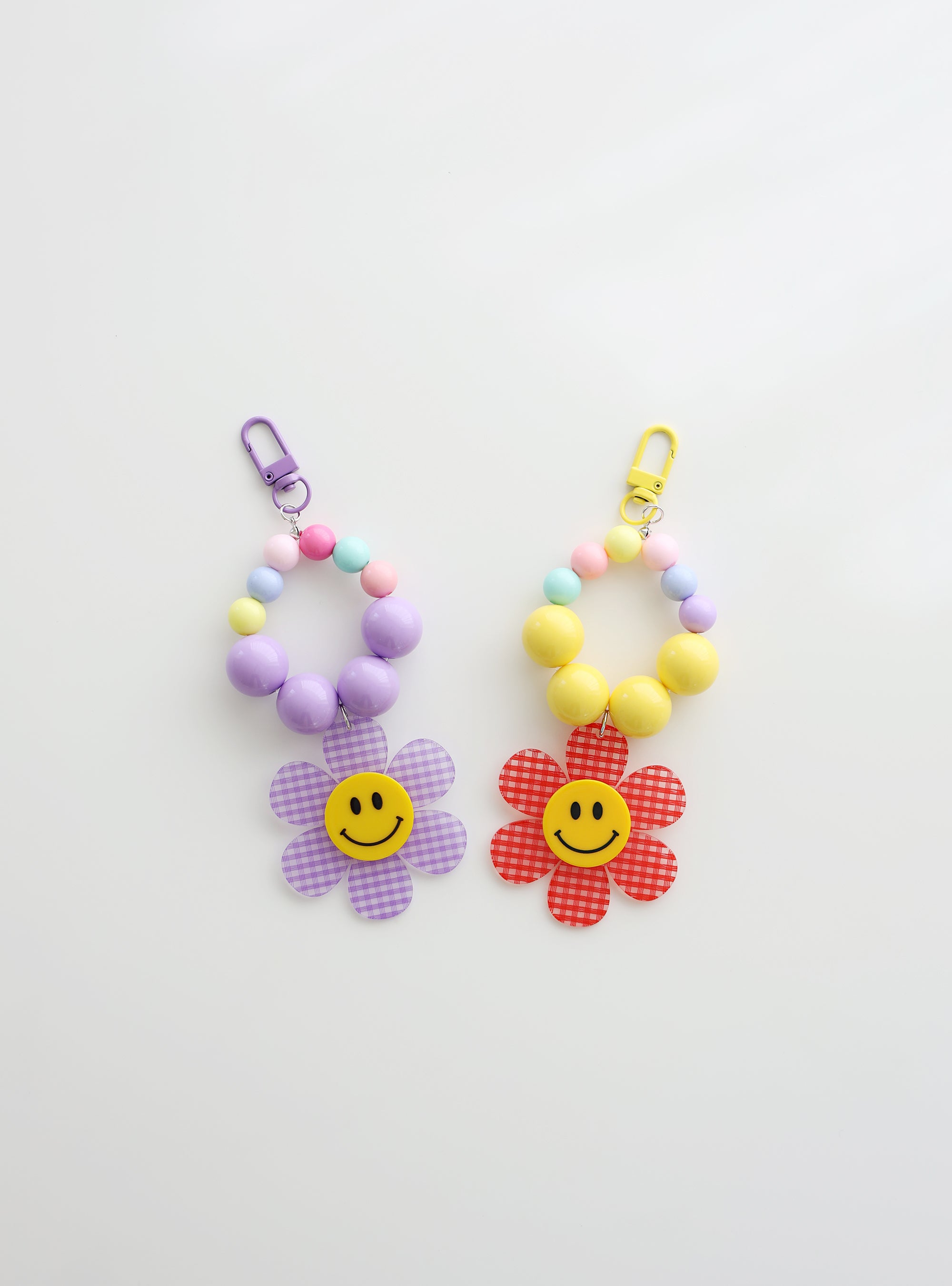 Two colorful beaded keychains with gingham flower smiley charms — one in soft purple and one in red — on a white background.