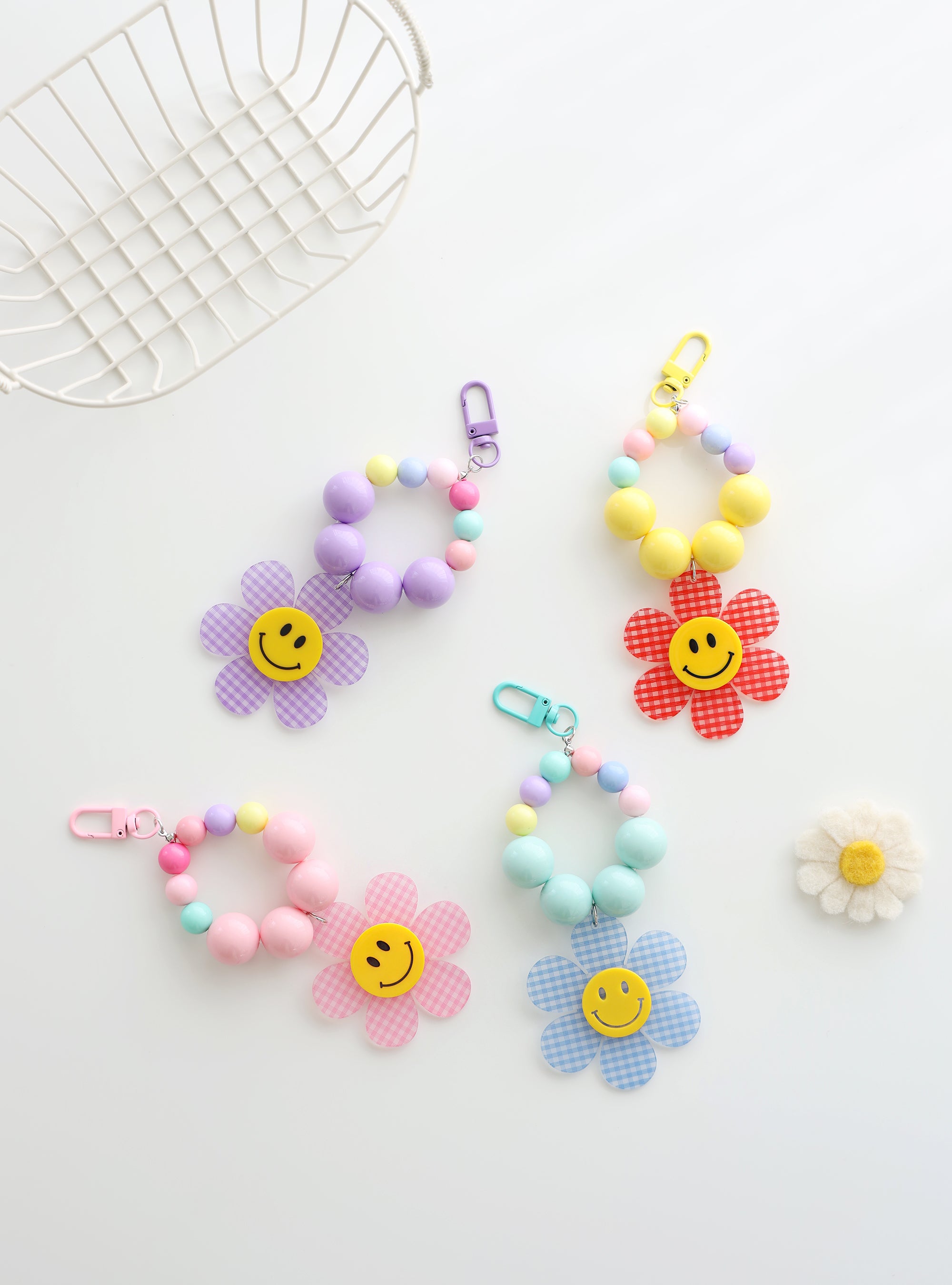 Four colorful beaded keychains with large pastel beads and playful gingham flower charms featuring smiley faces, displayed on a white background with white basket and a daisy decoration.