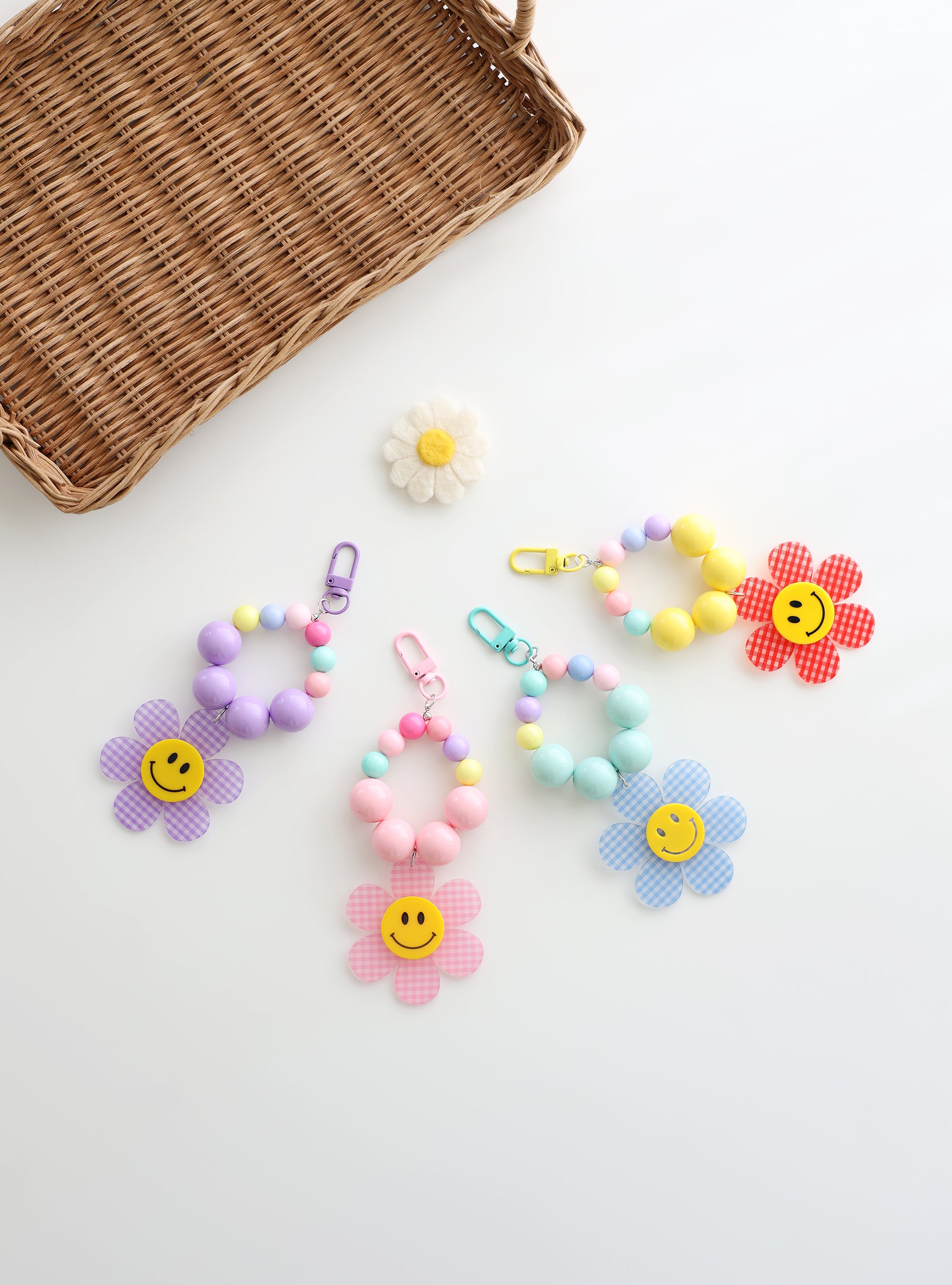 Four colorful beaded keychains with large pastel beads and playful gingham flower charms featuring smiley faces, displayed on a white background with basket and a daisy decoration.