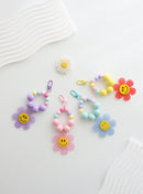 Four colorful beaded keychains with large pastel beads and playful gingham flower charms featuring smiley faces, displayed on a wavy white surface.
