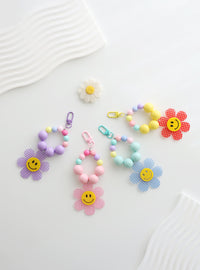 Four colorful beaded keychains with large pastel beads and playful gingham flower charms featuring smiley faces, displayed on a wavy white surface.