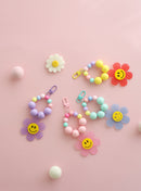 Four colorful beaded keychains with large pastel beads and playful gingham flower charms featuring smiley faces, displayed on a pink background with felt balls and a daisy decoration.