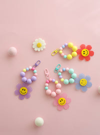 Four colorful beaded keychains with large pastel beads and playful gingham flower charms featuring smiley faces, displayed on a pink background with felt balls and a daisy decoration.