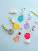 Six colorful smiley face keychains, each with round beads in playful pastel and bold colors, arranged on a light blue background with two small felt clouds and yellow pencils for a fun, cheerful display.