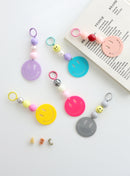 Six colorful smiley face keychains with playful bead accents in pastel and bold shades, displayed on a white surface with one keychain resting on a book.