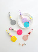 Six colorful smiley face keychains with playful bead accents in pastel and bold shades, displayed on a white surface with one keychain resting on a plate decorated with blue writing.