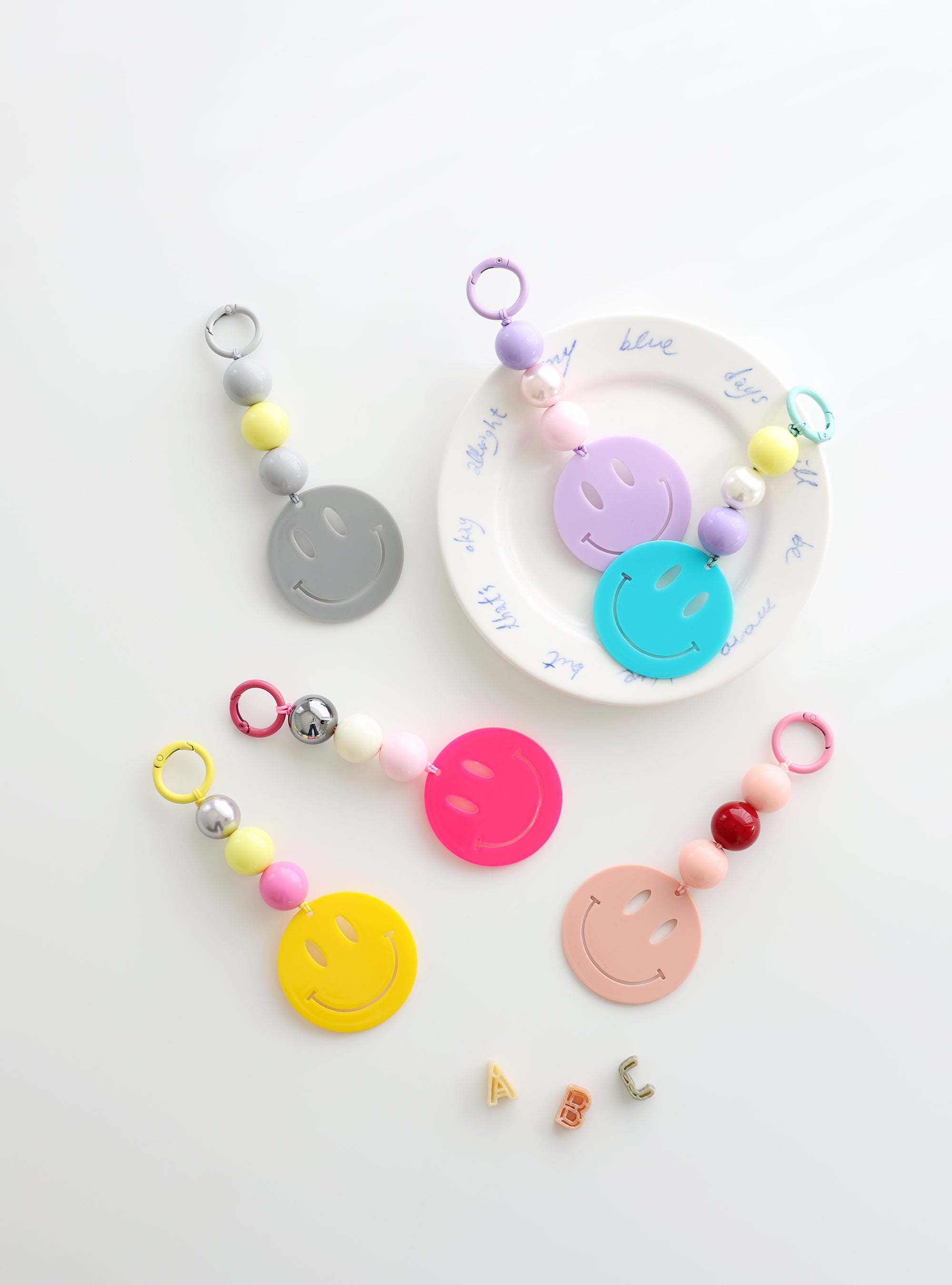 Six colorful smiley face keychains with playful bead accents in pastel and bold shades, displayed on a white surface with one keychain resting on a plate decorated with blue writing.
