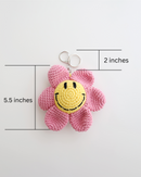 A crocheted flower keychain with soft pink petals and a bright yellow smiley face center is displayed. It features a silver keyring at the top for easy attachment to bags, keys, or pouches. The picture includes size measurement of 5.5inches in height, 2 inches of keyring height totaled 7.5inches in height. 
