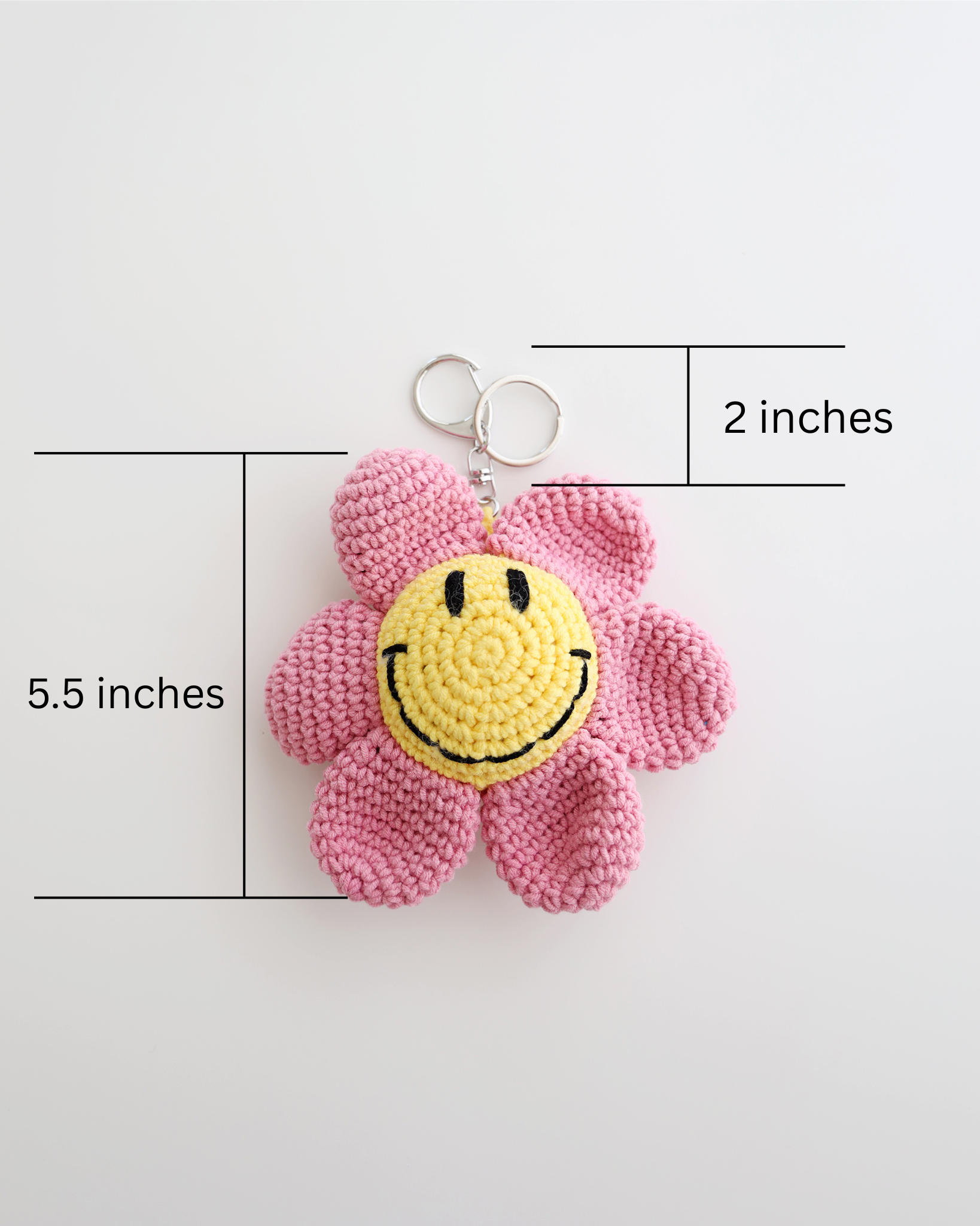 A crocheted flower keychain with soft pink petals and a bright yellow smiley face center is displayed. It features a silver keyring at the top for easy attachment to bags, keys, or pouches. The picture includes size measurement of 5.5inches in height, 2 inches of keyring height totaled 7.5inches in height. 