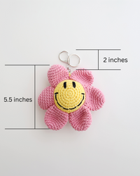 A crocheted flower keychain with soft pink petals and a bright yellow smiley face center is displayed. It features a silver keyring at the top for easy attachment to bags, keys, or pouches. The picture includes size measurement of 5.5inches in height, 2 inches of keyring height totaled 7.5inches in height. 