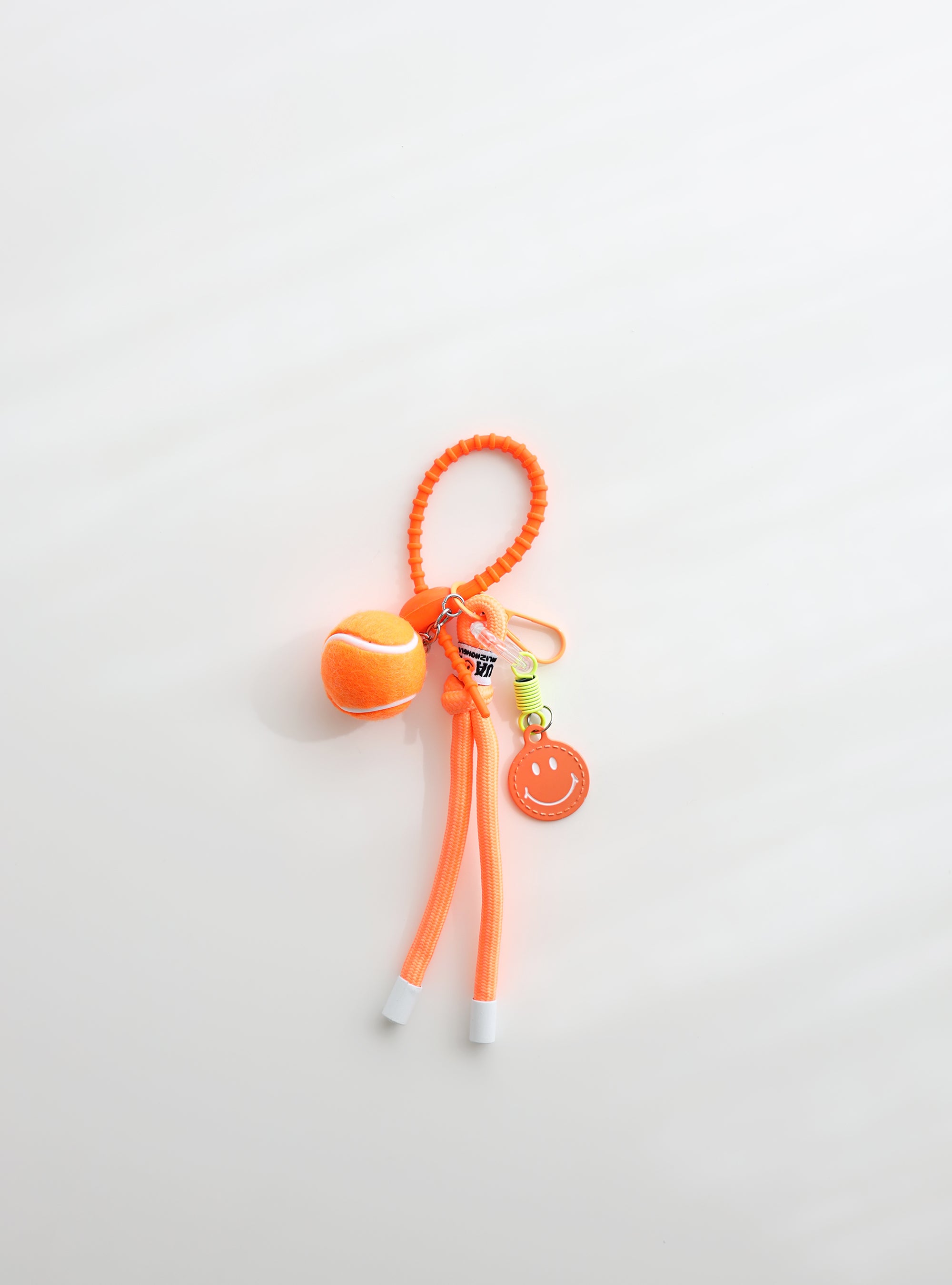A bright orange tennis ball keychain featuring a mini tennis ball charm, a orange smiley face tag, and an adjustable loop strap for easy attachment, displayed on a clean white background.