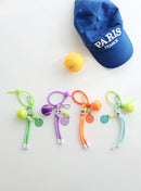 Four colorful tennis ball keychains in green, purple, orange, and mint, each with a mini tennis ball charm, smiley face tag, and adjustable loop strap, displayed on a white surface with a blue “Paris France” cap and a yellow tennis ball in the background.