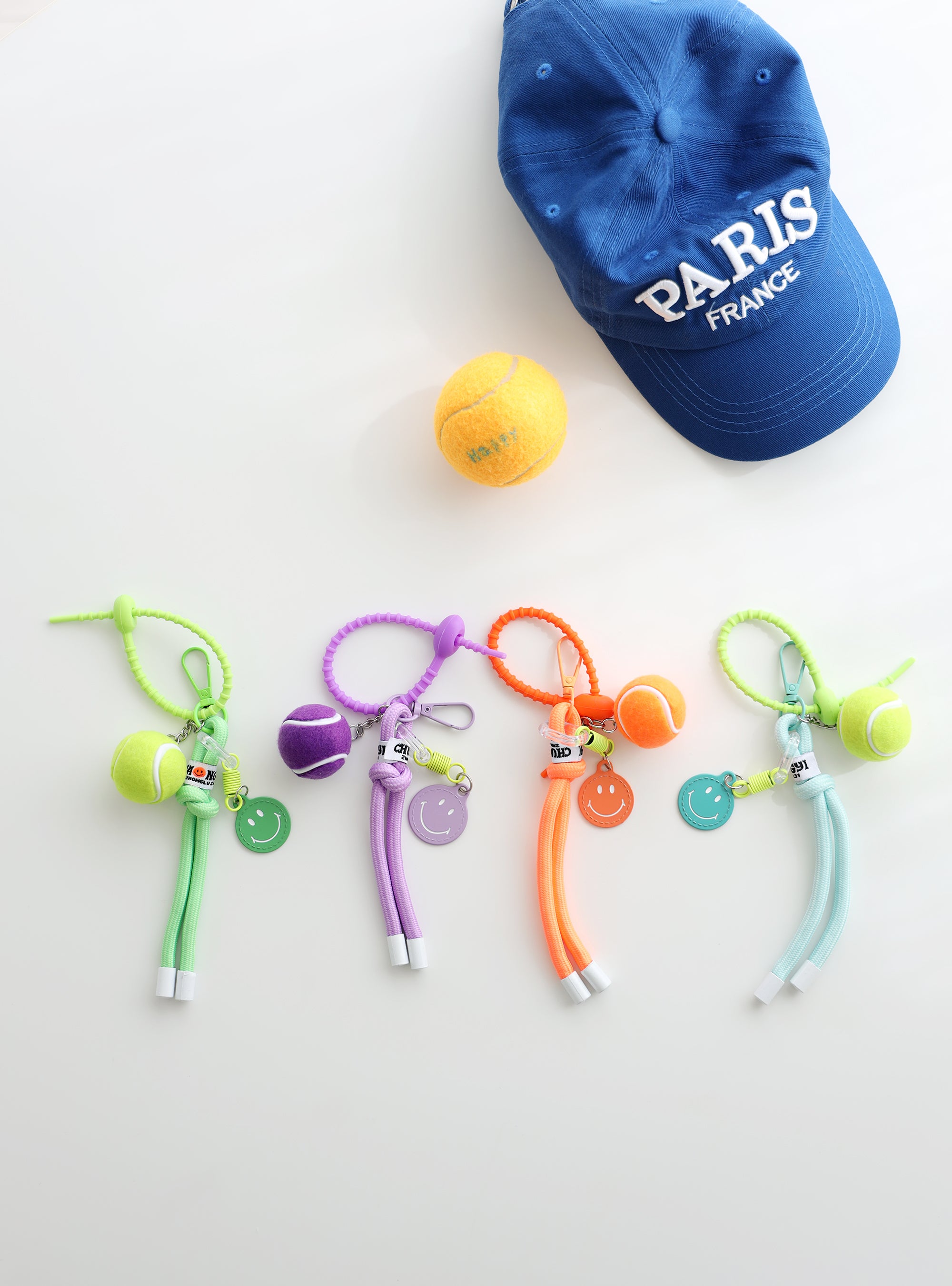 Four colorful tennis ball keychains in green, purple, orange, and mint, each with a mini tennis ball charm, smiley face tag, and adjustable loop strap, displayed on a white surface with a blue “Paris France” cap and a yellow tennis ball in the background.