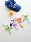Four colorful tennis ball keychains in green, purple, orange, and mint, each with a mini tennis ball charm, smiley face tag, and adjustable loop strap, displayed on a white surface with a blue “Paris France” cap and a yellow tennis ball in the background.