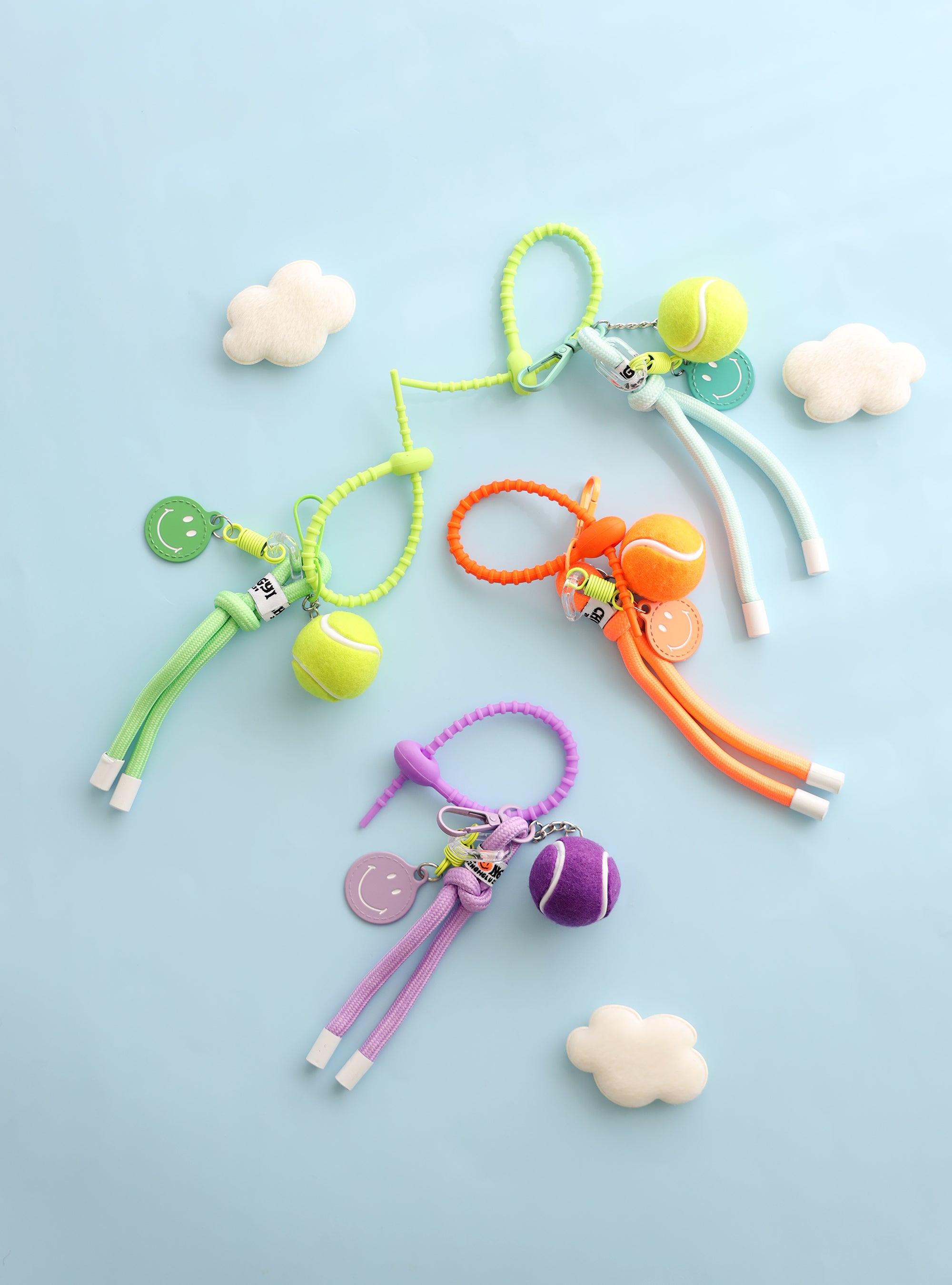 Four colorful tennis ball keychains in green, mint, orange, and purple, each featuring a mini tennis ball charm, a smiley face tag, and a loop strap for easy attachment, displayed on a light blue background with small felt clouds.