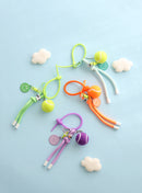 Four colorful tennis ball keychains in green, mint, orange, and purple, each featuring a mini tennis ball charm, a smiley face tag, and a loop strap for easy attachment, displayed on a light blue background with small felt clouds.