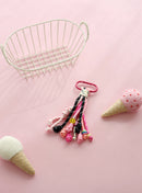 A playful keychain accessory featuring multiple knotted ropes in shades of pink, black, and white, with a small yellow smiley charm and a bright pink clasp, displayed on a pink background with felt ice cream cone props.