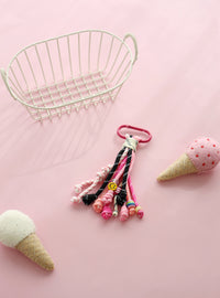 A playful keychain accessory featuring multiple knotted ropes in shades of pink, black, and white, with a small yellow smiley charm and a bright pink clasp, displayed on a pink background with felt ice cream cone props.