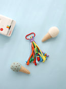 A playful keychain accessory featuring multiple knotted ropes in shades of sky blue, orange, green, and yellow, with a small yellow smiley charm and a bright red clasp, displayed on a sky blue background with felt ice cream cone props.