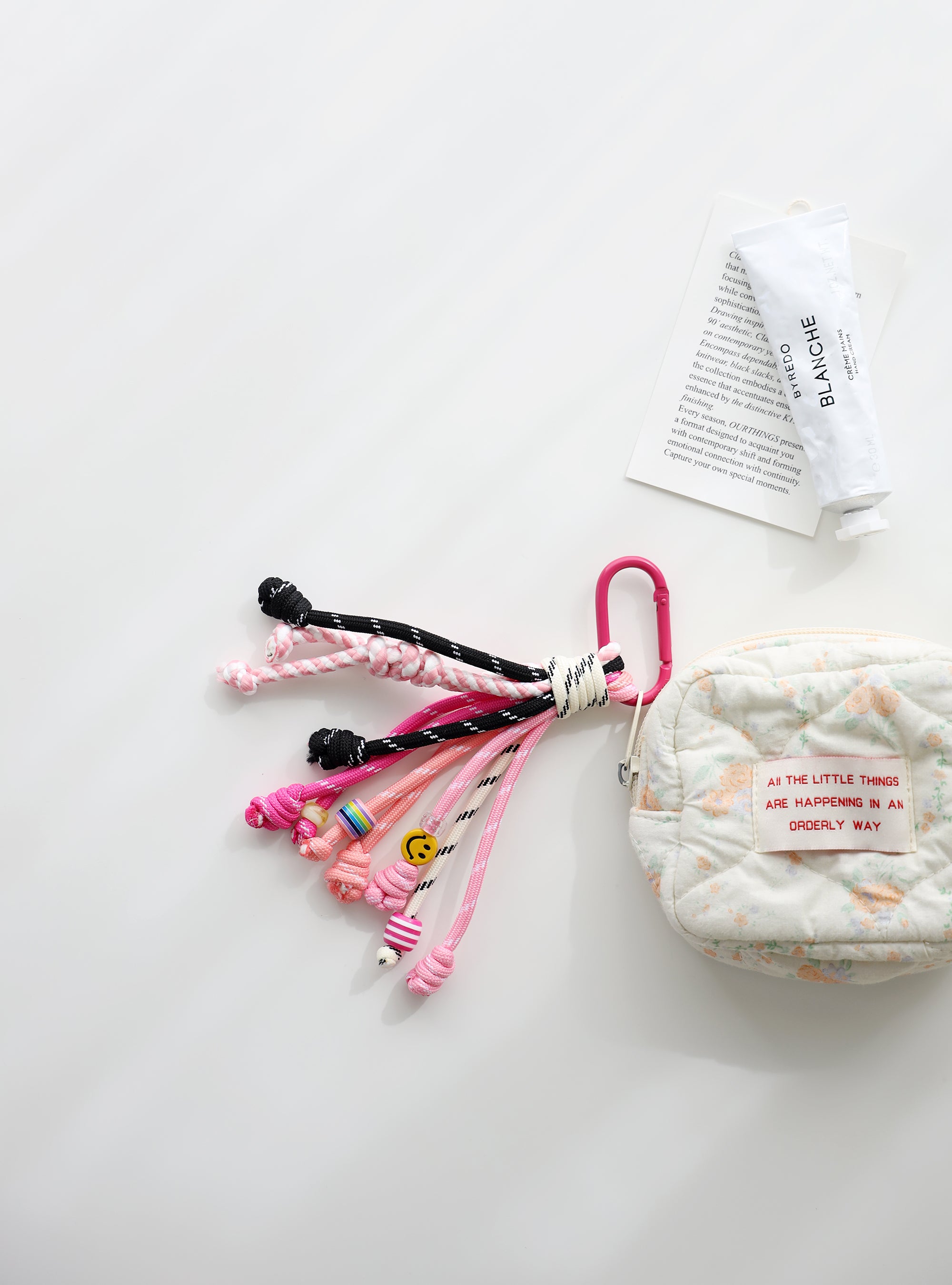 A playful multi-rope keychain featuring knotted cords in pink, black, and white with colorful beads and a small yellow smiley charm, attached to a pink clasp and shown clipped to a quilted pouch on a white background.
