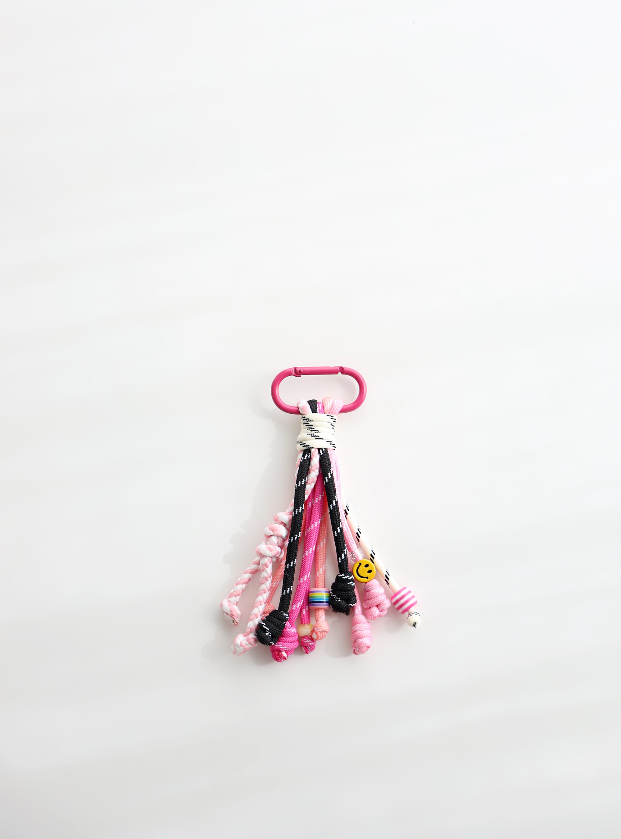 A playful multi-rope keychain featuring knotted cords in pink, black, and white with colorful beads and a small yellow smiley charm, attached to a pink clasp on a white background.