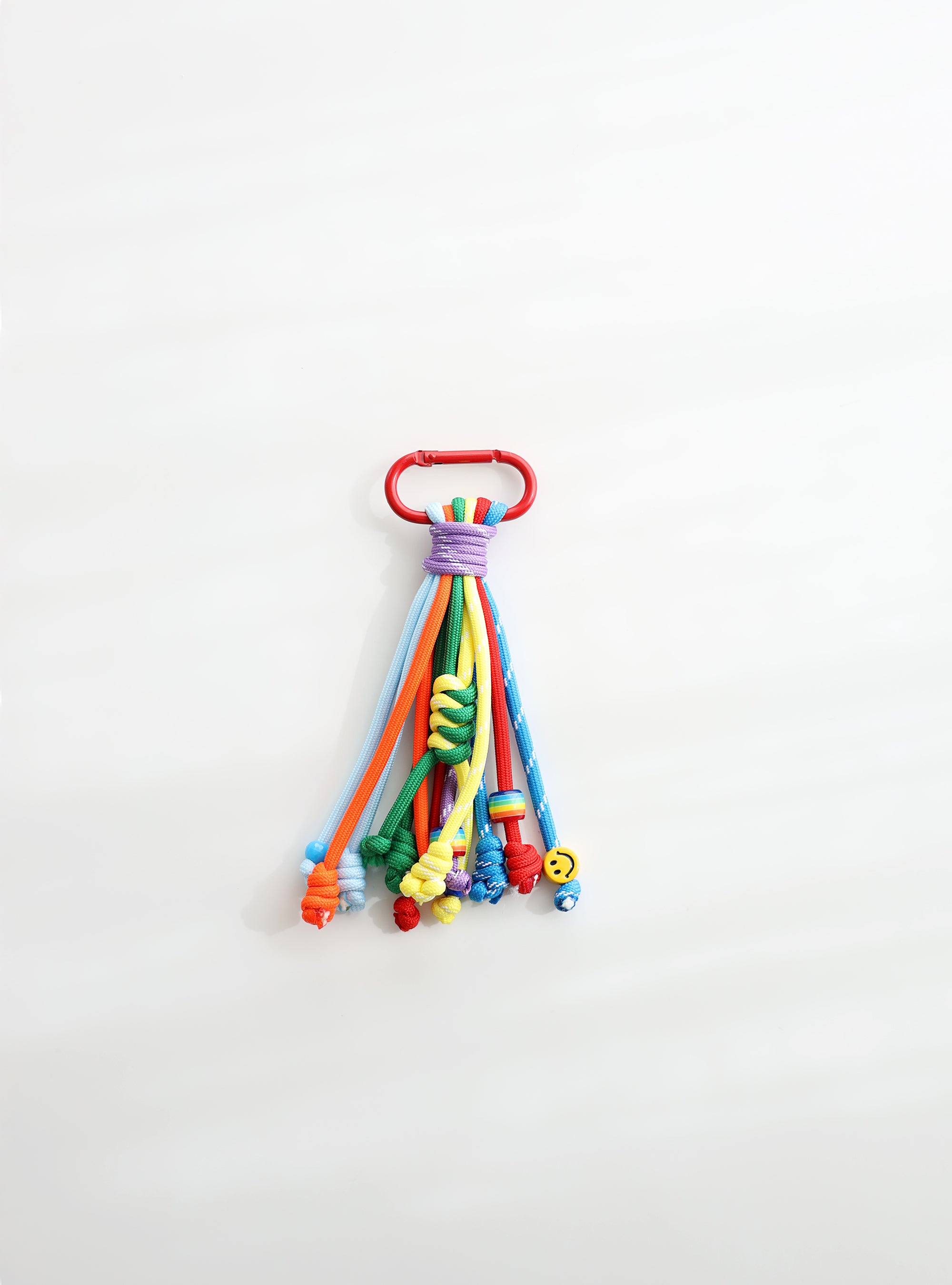 A playful multi-rope keychain featuring knotted cords in orange, sky blue, green and yellow with colorful beads and a small yellow smiley charm, attached to a red clasp on a white background.
