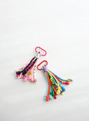 Two colorful rope keychains with multiple knotted strands — one in pink, black, and white tones, the other in bright rainbow colors — each with playful bead details, small smiley charms, and metal clasps for easy attachment.
