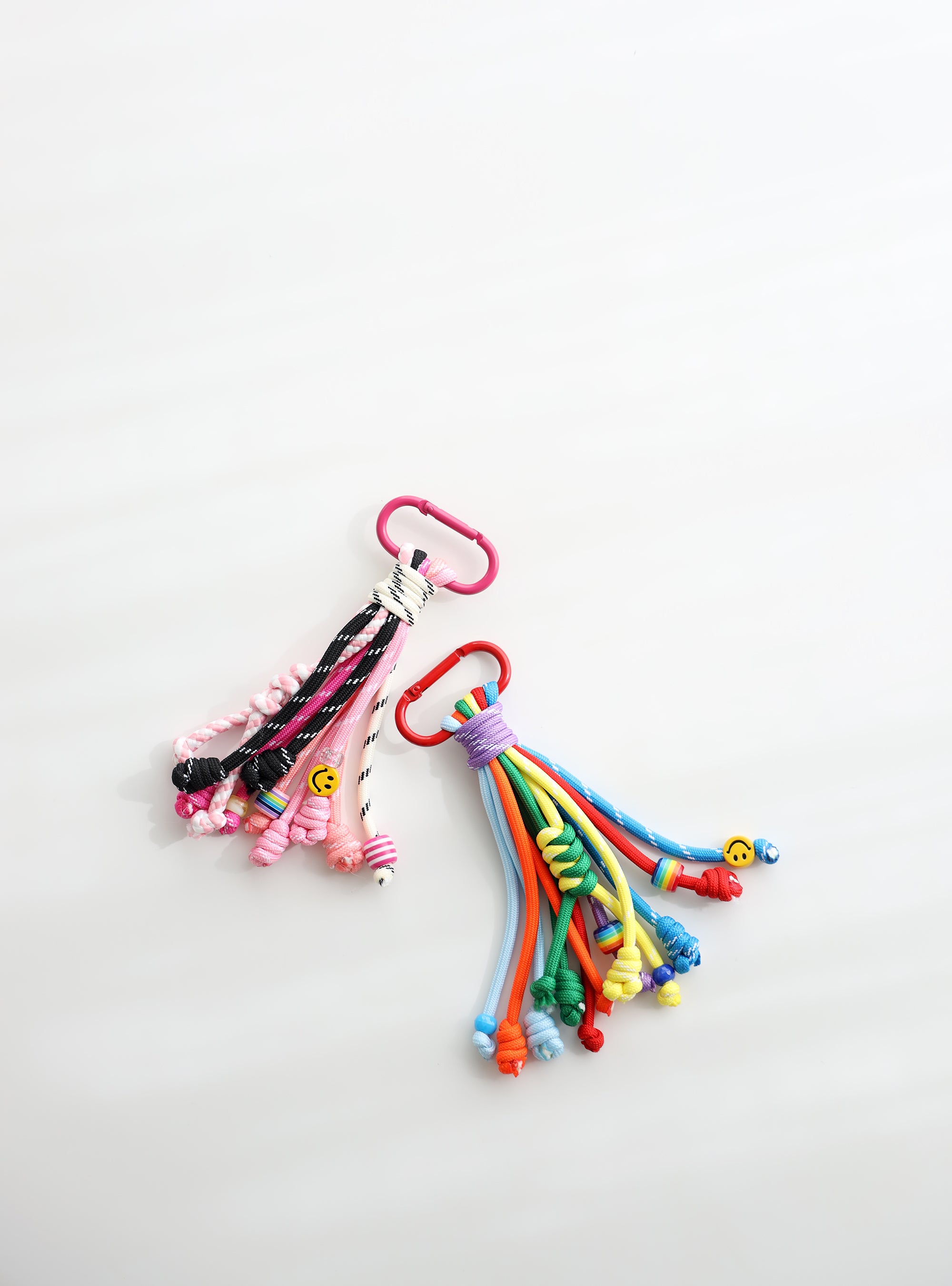 Two colorful rope keychains with multiple knotted strands — one in pink, black, and white tones, the other in bright rainbow colors — each with playful bead details, small smiley charms, and metal clasps for easy attachment.