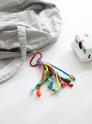 A playful multi-rope keychain featuring knotted cords in orange, sky blue, green and yellow with colorful beads and a small yellow smiley charm, attached to a red clasp and shown clipped to a grey bag on a white background.