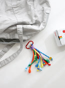 A playful multi-rope keychain featuring knotted cords in orange, sky blue, green and yellow with colorful beads and a small yellow smiley charm, attached to a red clasp and shown clipped to a grey bag on a white background.