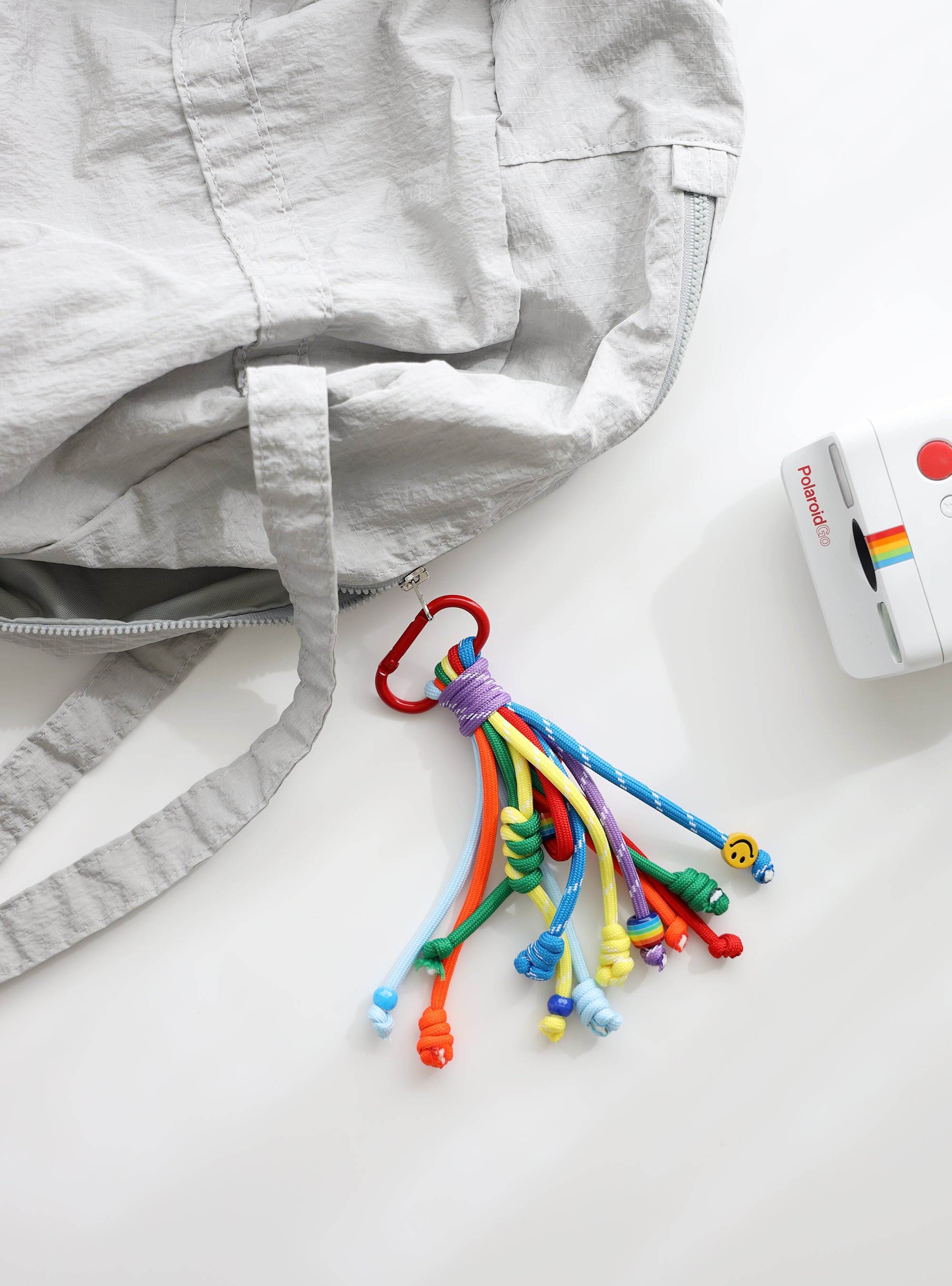 A playful multi-rope keychain featuring knotted cords in orange, sky blue, green and yellow with colorful beads and a small yellow smiley charm, attached to a red clasp and shown clipped to a grey bag on a white background.